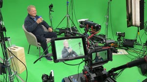 An old man sat in a chair on a green screen set surrounded by cameras