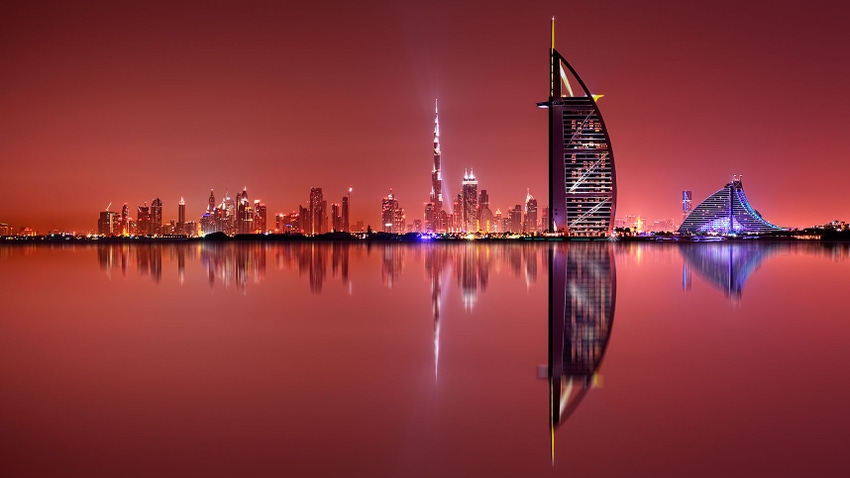 Dubai skyline reflected onto water. The setting sun casts a red glow.