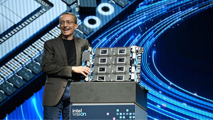 A man in a grey jacket, blue jeans and a black t-shirt holding a stack of semiconductors up on stage in front of a blue background