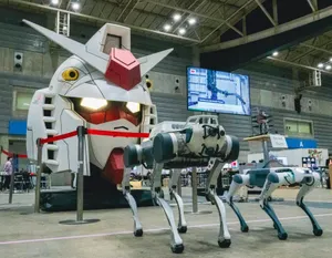 Two quadrupedal robots stand in front of a giant Gundamn head 