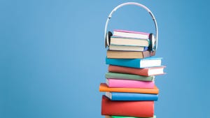 A pile of multicolored books stacked up high against a blue background. A pair of white headphones sits atop them. 
