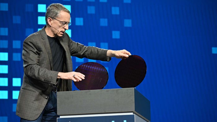 A man in a grey jacket, blue jeans and a black t-shirt holding up two circular semiconductors on stage in front of a blue background