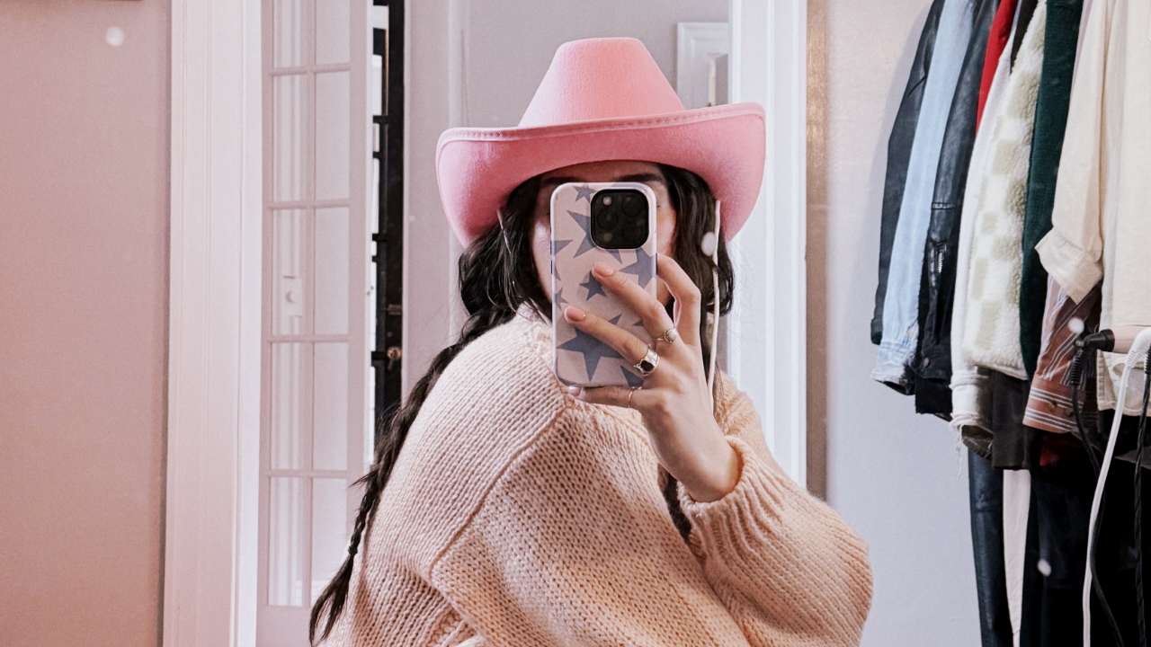 A girl in a pink cowboy hat is taking a picture of herself with her cell phone in the mirror.