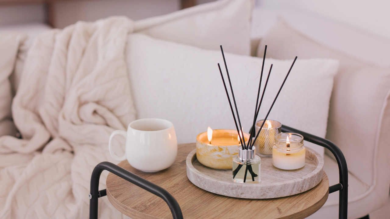 Un arrangement chaleureux comprenant une petite table sur laquelle reposent une tasse blanche, des chandelles allumées et des diffuseurs en roseau, placés devant des coussins beiges moelleux et une couverture en tricot.
