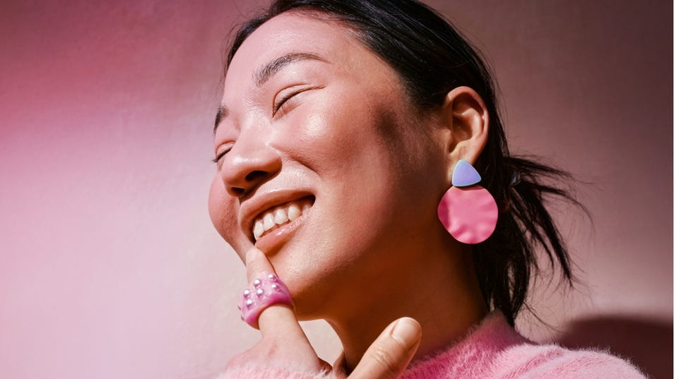 A woman in a pink sweater laughs with her hand near her face, eyes closed in a joyful expression.