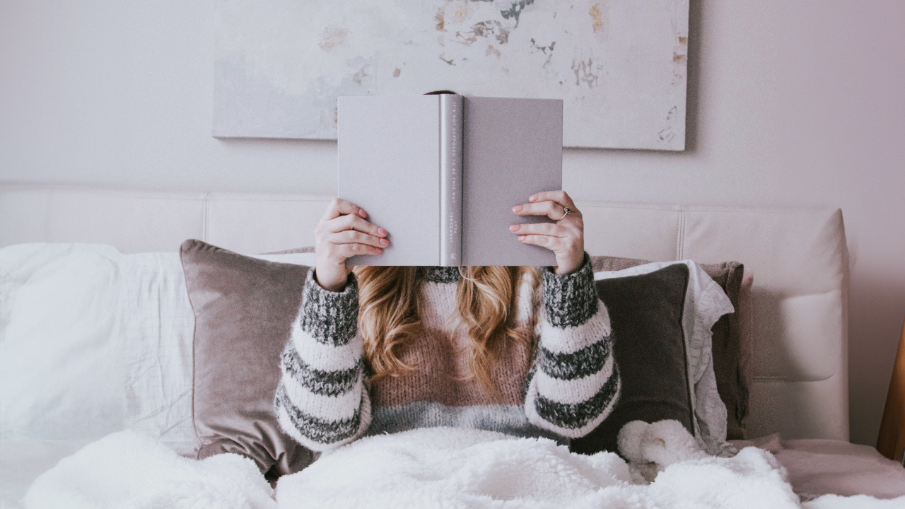 Une fille assise dans son lit porte un chandail douillet et lit un livre.