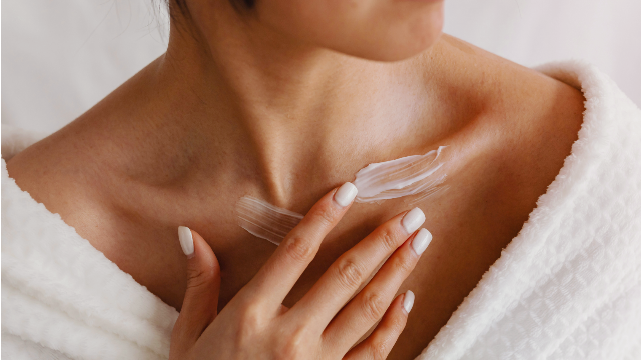 A close-up of a woman in a white robe applying a moisturizing lotion to her collarbone area