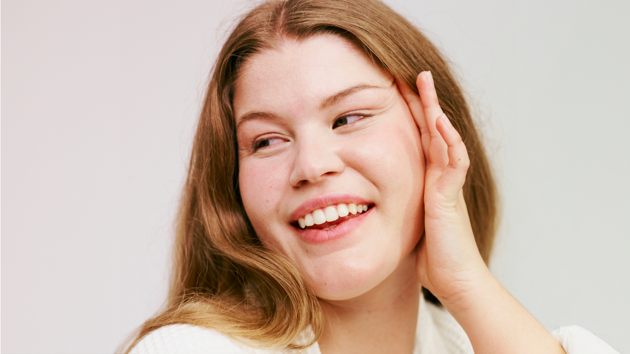 A smiling woman with long, wavy hair touching her face gently, exuding confidence and joy in soft natural lighting.