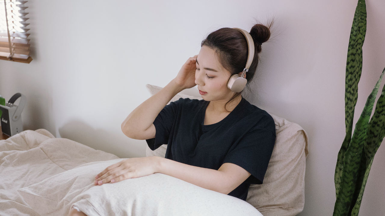 Une femme portant des écouteurs, assise sur un lit, l’air calme, dans une pièce à l’éclairage tamisé.