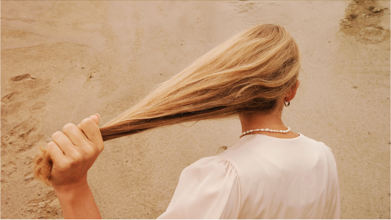 A woman stands with her back to the camera, holding out her blonde hair with one hand.