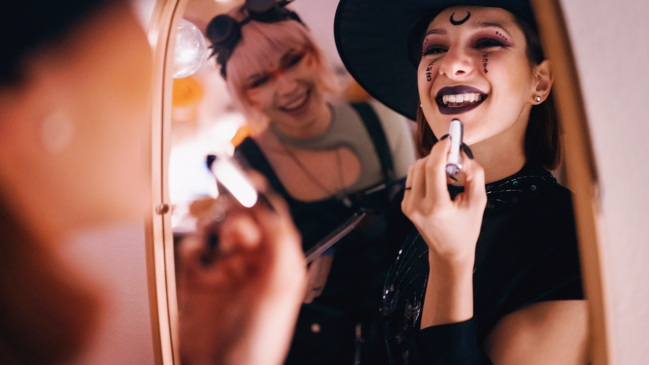 Two women are getting dressed up for Halloween, doing their make-up together, and looking in a mirror.
