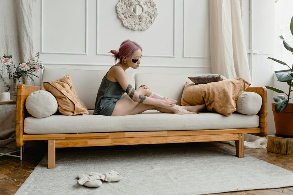 Une femme aux cheveux roses et au bras tatoué d’un serpent est assise sur un sofa et s’applique des bandes de cire sur les jambes.