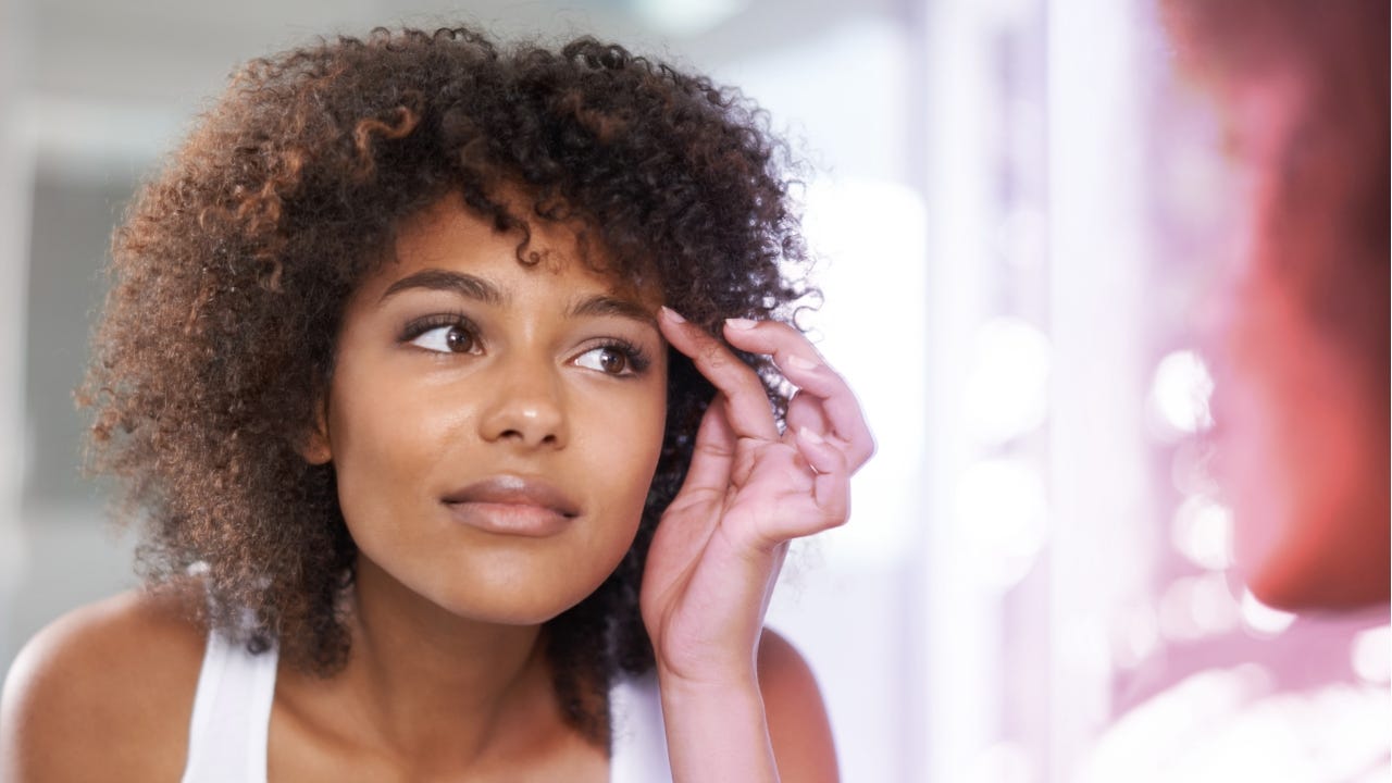 A young woman with curly hair examines her reflection in the mirror while gently touching her eyebrow.