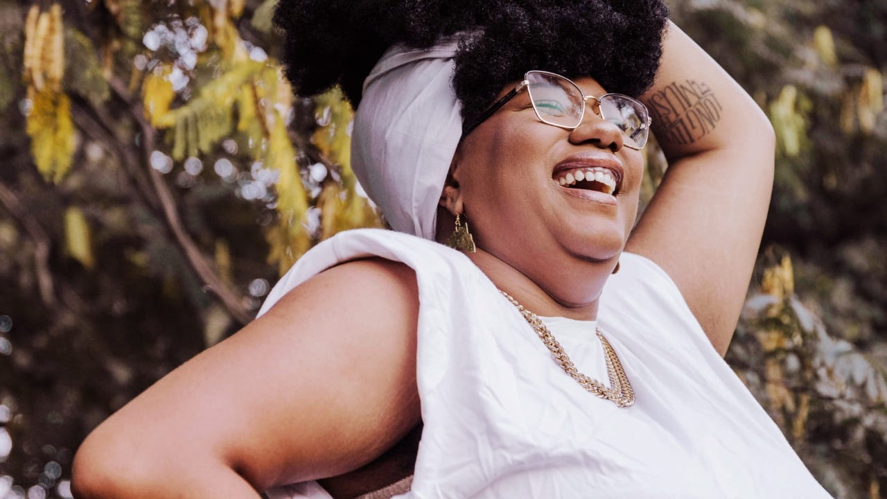 Une femme portant des lunettes et vêtue de blanc rit en plein air, dévoilant une aisselle lisse.