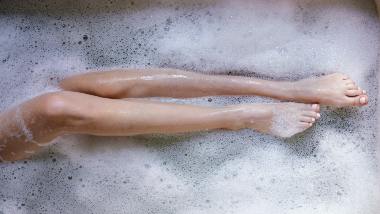 A pair of smooth, hairless legs stretched out in a bathtub filled with frothy, white bubbles.