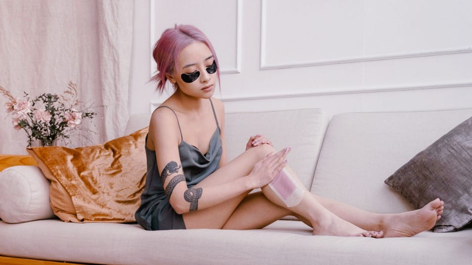 Une femme aux cheveux roses et au bras tatoué d’un serpent est assise sur un sofa tout en s’appliquant des bandes de cire sur les jambes.