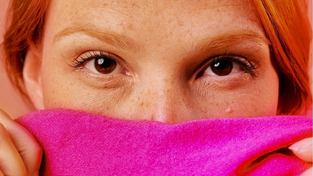 A close-up of a woman with red hair and freckles gazing directly into the camera while partially covering her face with a bright pink sweater.
