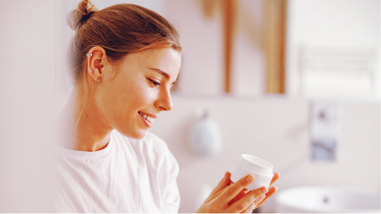 A woman in white shirt reading the label of a face cream container.
