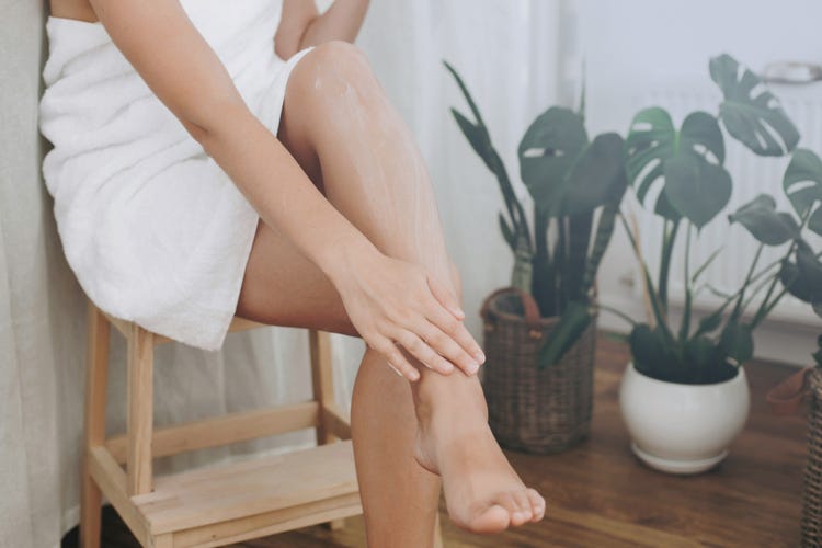Une femme enveloppée dans une serviette est assise sur un tabouret à côté de deux plantes et caresse ses jambes lisses.