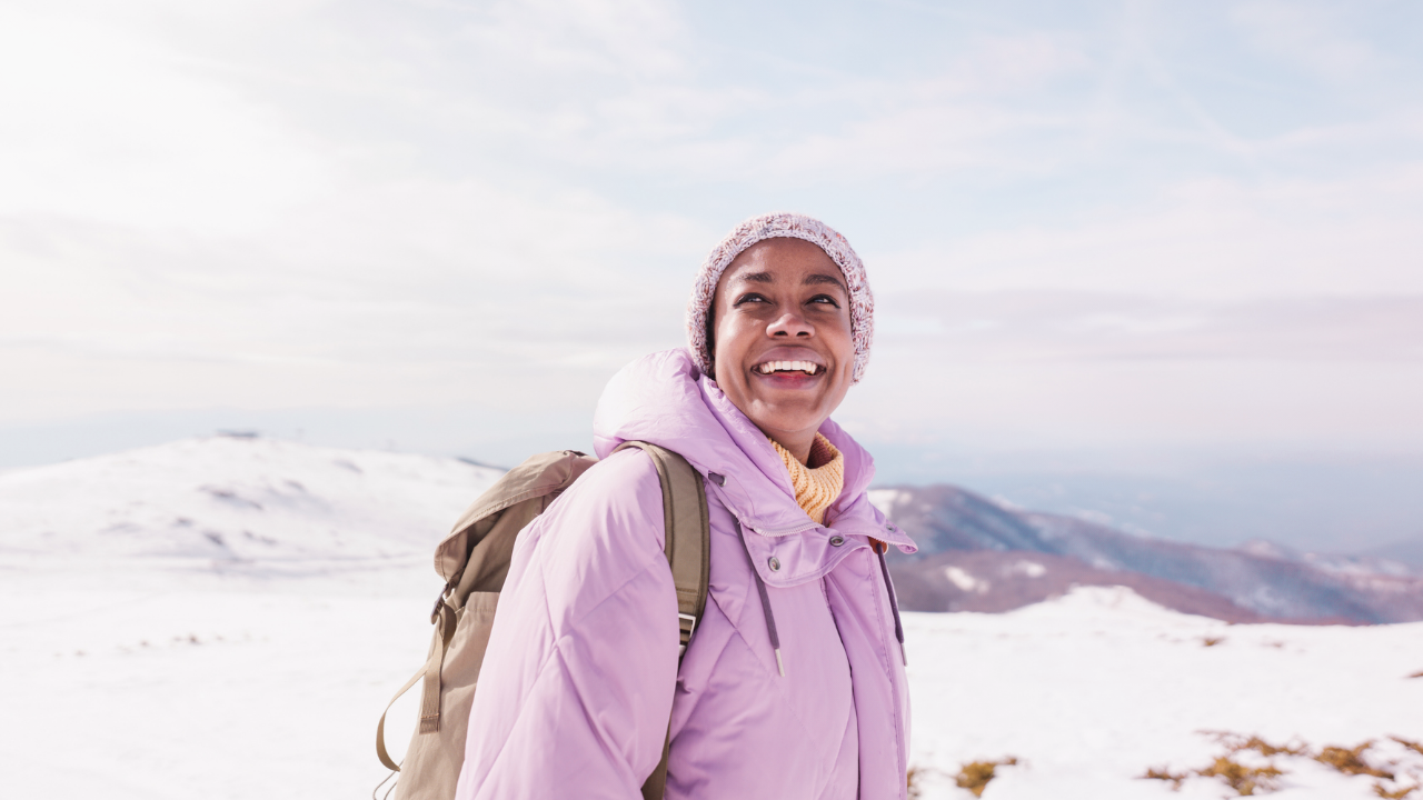 Une femme vêtue d’un manteau et d’une tuque de couleur lavande, l’air joyeux, qui explore une montagne enneigée.