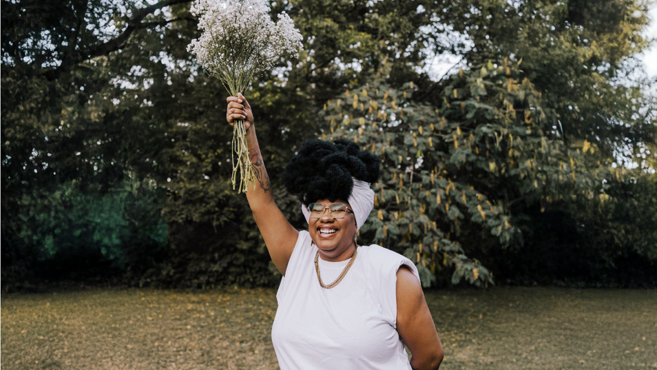 Une femme en camisole, debout sur une pelouse, tient un bouquet de fleurs blanches dans les airs.