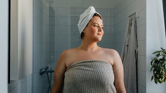 Dans une salle de bain, une femme confiante, les cheveux et le corps enveloppés dans une serviette, s’apprête à commencer sa douche complète. 