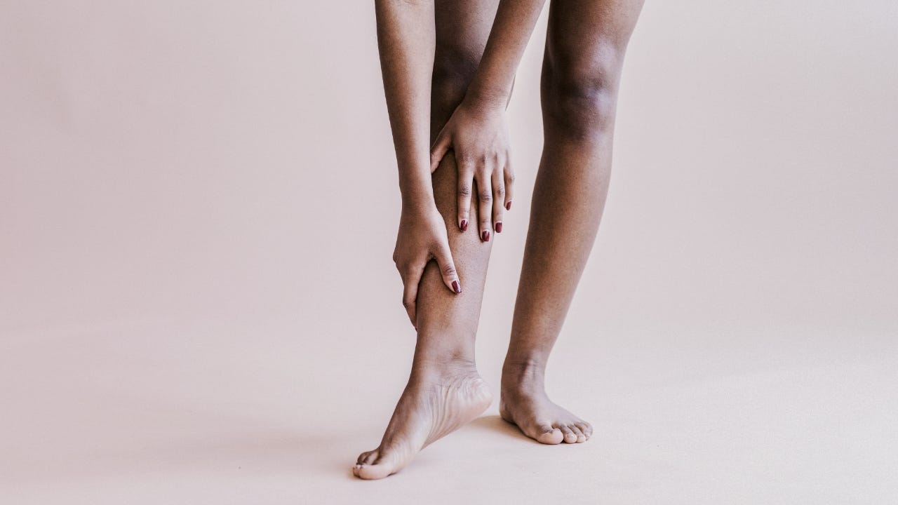 Une personne aux jambes lisses touche doucement son mollet sur un fond neutre.