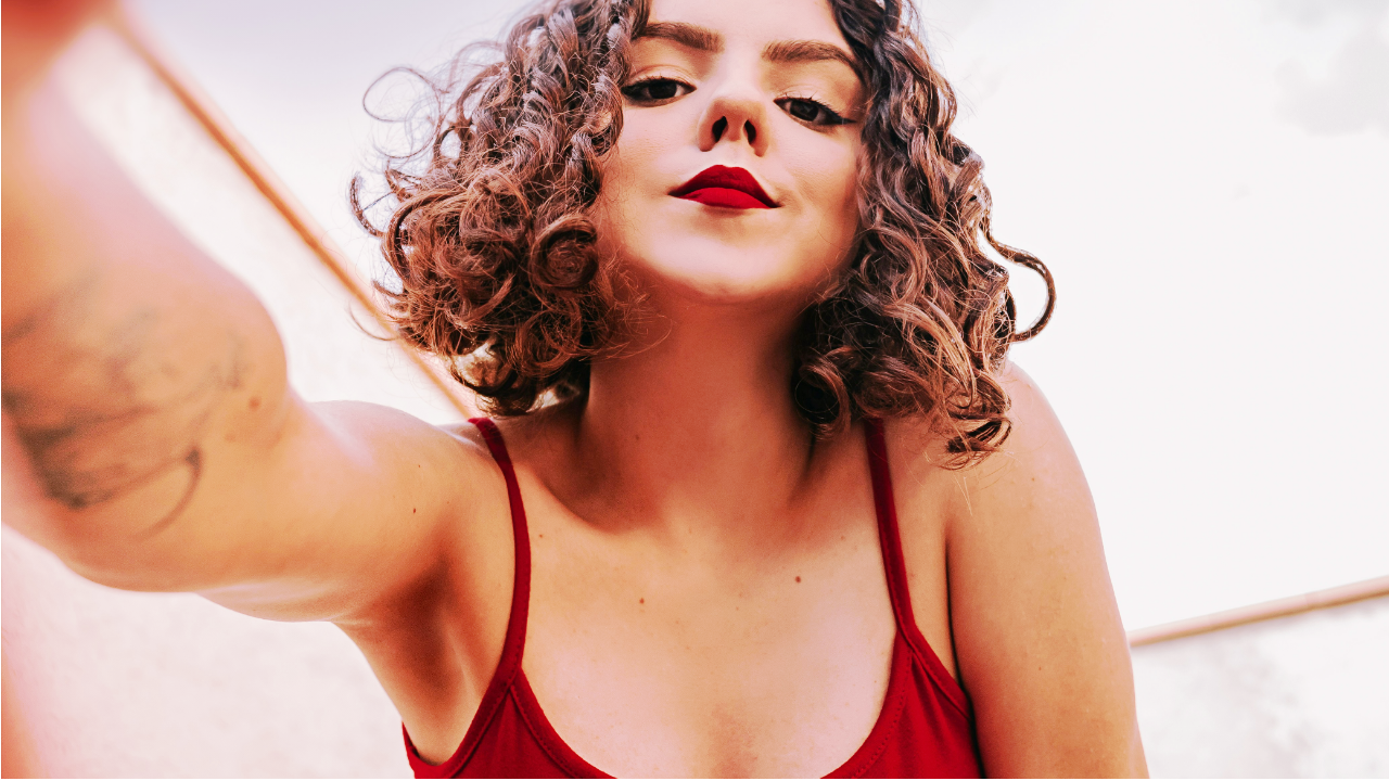 Une femme aux cheveux courts et frisés, portant du rouge à lèvres et une camisole rouges, regarde vers le bas, directement dans l’objectif.