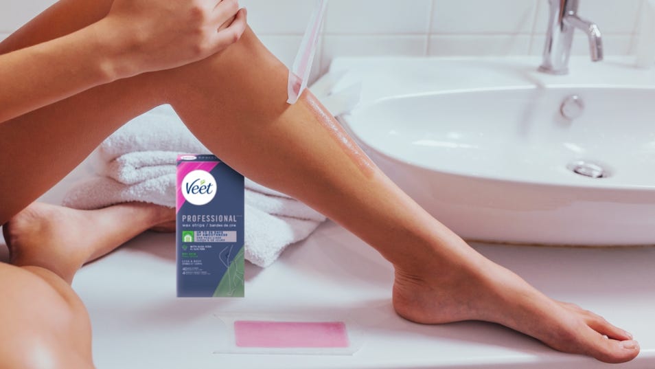 Une femme, assise sur un comptoir de salle de bain, retire une bande de cire rose Veet Professional de sa jambe. L’emballage du produit est visible à proximité.