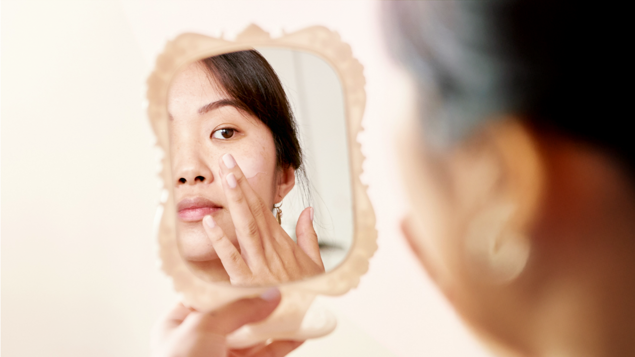 A close-up of a woman using her fingertips to apply cream to her cheek, captured through the reflection of a decorative mirror.