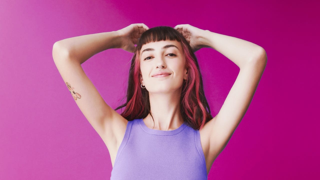 A confident woman with smooth underarms posing with hands behind their head against a vibrant pink background.