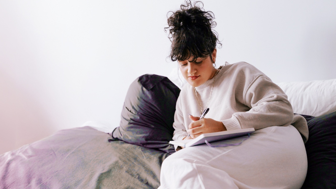 A young woman in a sweatshirt lounging on her bed, writing in a journal with a thoughtful expression.