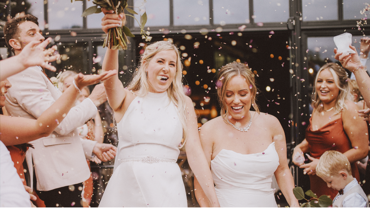 Two brides celebrating joyfully as guests toss flower petals.