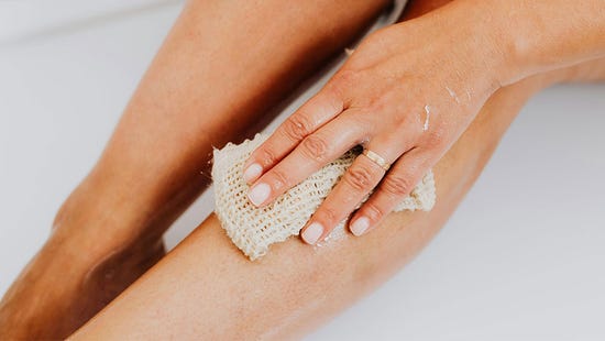 Une personne assise dans la baignoire exfolie ses jambes avec un louffa dans la main.