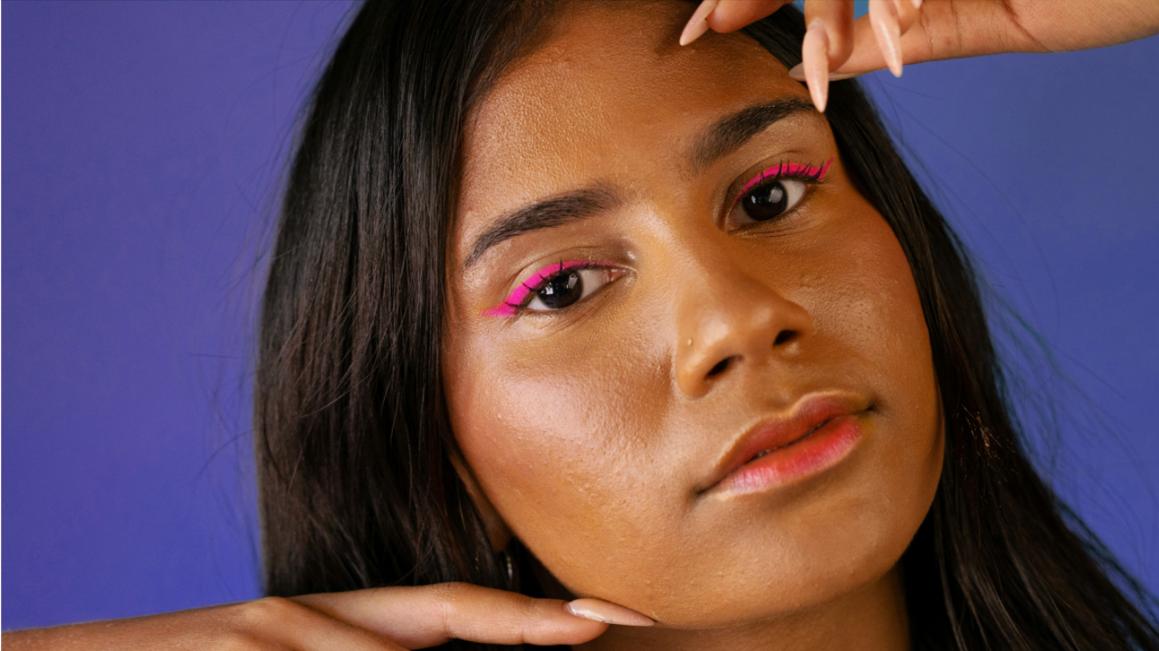 close-up portrait of a woman with radiant skin, striking pink makeup, and sleek dark hair, set against a blue backdrop.