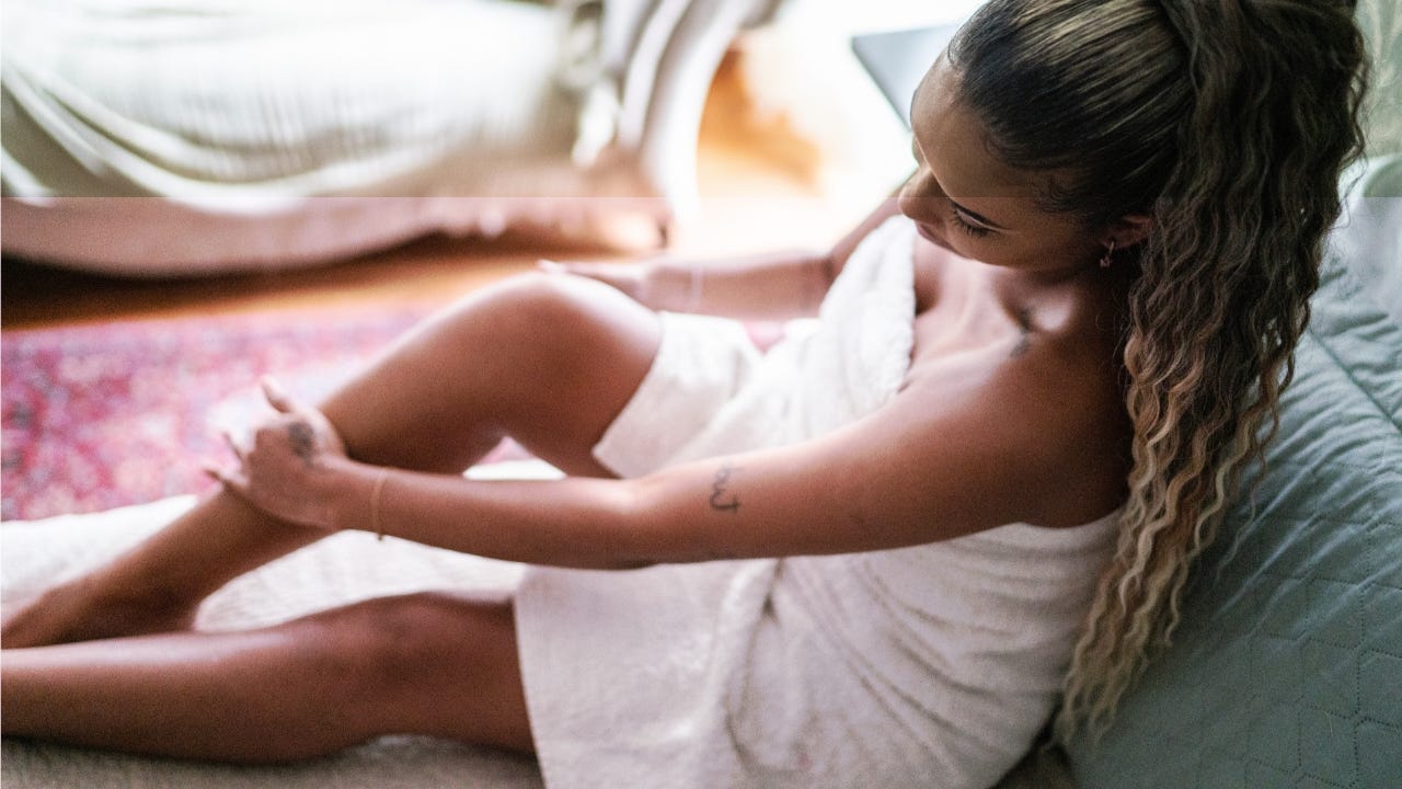 Une femme enveloppée d’une serviette blanche est assise au sol, caressant doucement ses jambes lisses dans une chambre à l’éclairage tamisé.