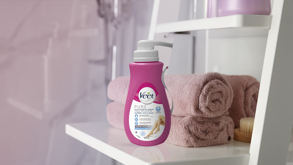 Un contenant de crème dépilatoire Veet PURE placé sur une tablette blanche, à côté de serviettes roses soigneusement enroulées et d’une brosse pour le corps dans une salle de bain moderne.