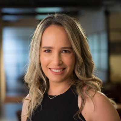Shira Shamban, CEO and Co-founder of Solvo, has long wavy blonde hair. She's wearing a black shirt and delicate gold necklace.