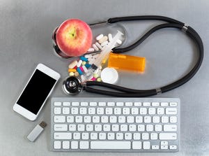 An apple, a computer keyboard, a stethoscope, and a bottle of pills on a grey table.