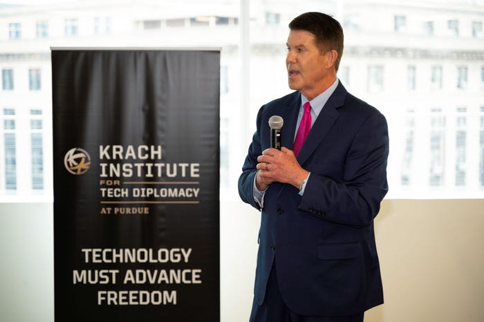 Keith Krach talks into a microphone in front of a banner for his Krach Institute for Tech Diplomacy