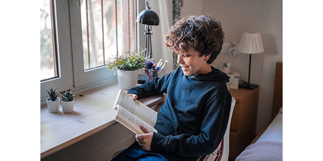 A smiling boy wearing a black hoodie is reading a book at his bedroom desk