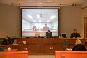 A courtroom with several judges behind the bench and a screen projection showing two people in another court room