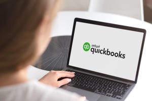 Image shows a woman with blonde hair from behind sitting at a laptop with an open screen showing the words "intuit quickbooks"