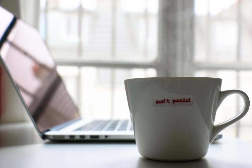 A white mug with red letters saying don't panic on a table with a laptop