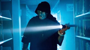 A man wearing a black hoodie and a silver mask walking in the dark between two bookshelves holding a flashlight A man wearing a black hoodie and a silver mask walking in the dark between two bookshelves holding a flashlight