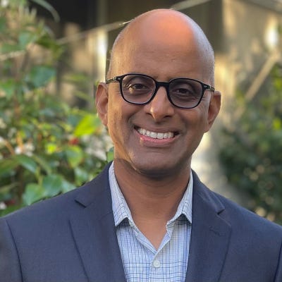 Raghu Yeluri has a shaved head and wears dark-framed glasses, an open-neck oxford, and a blue blazer in front of shrubbery