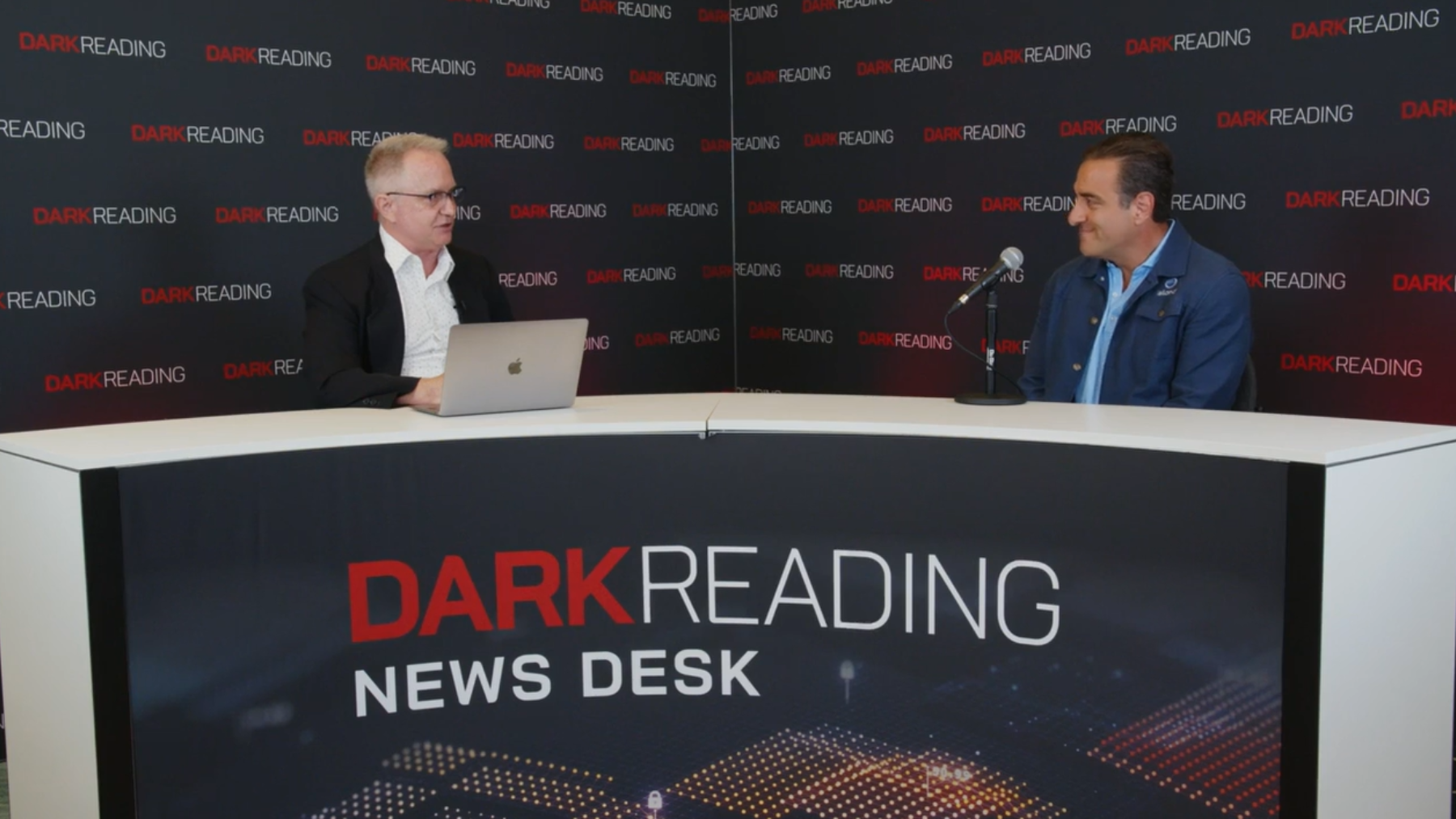  Two people talking at a news desk against a dark backdrop with "Dark Reading" written on it.