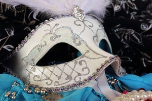 White masquerade ball mask adorned with glitter and rhinestons surrounded by party beads, bangles and fabrics