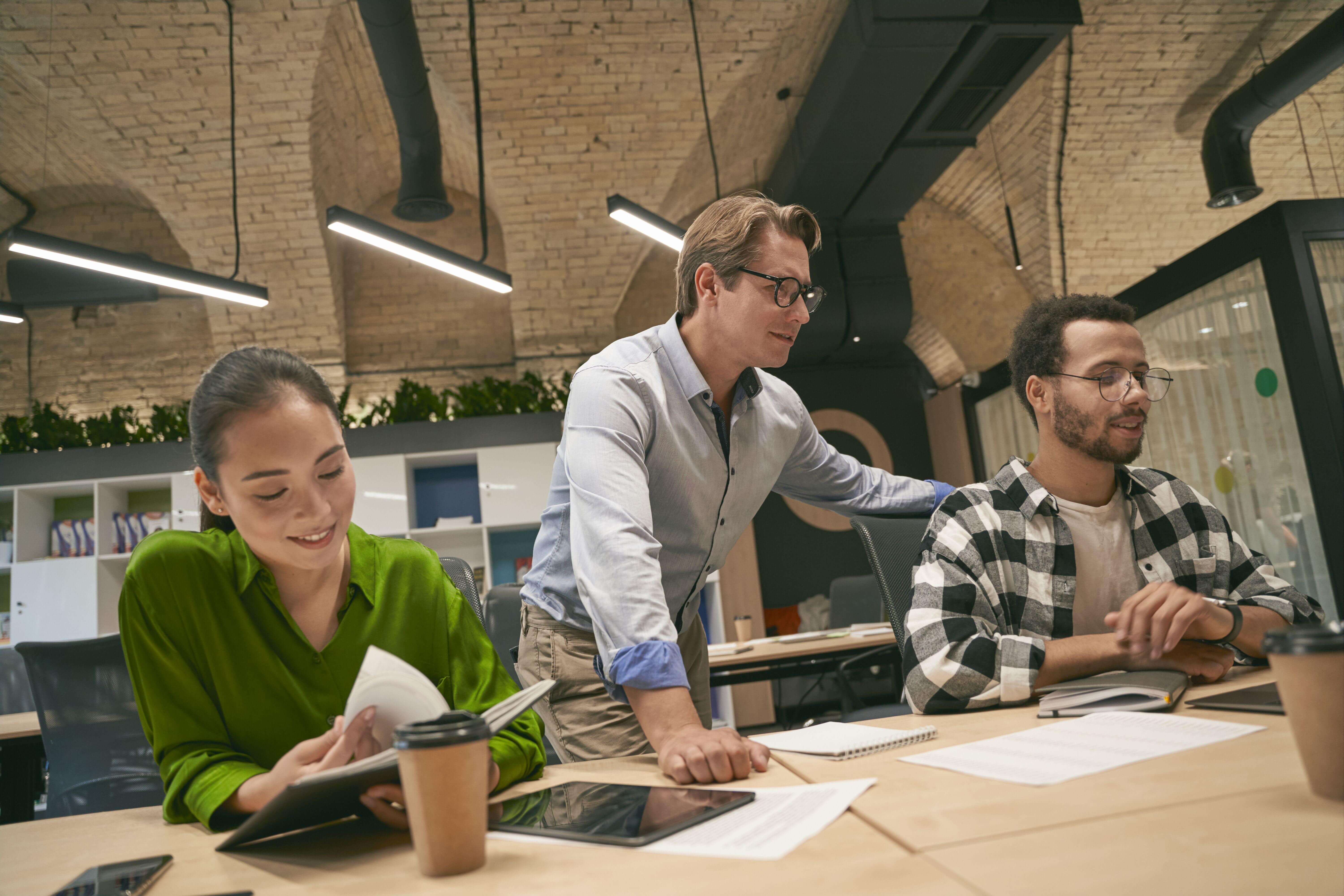 Man standing next to two seated coworkers who are working on a project together. They all look happy.
