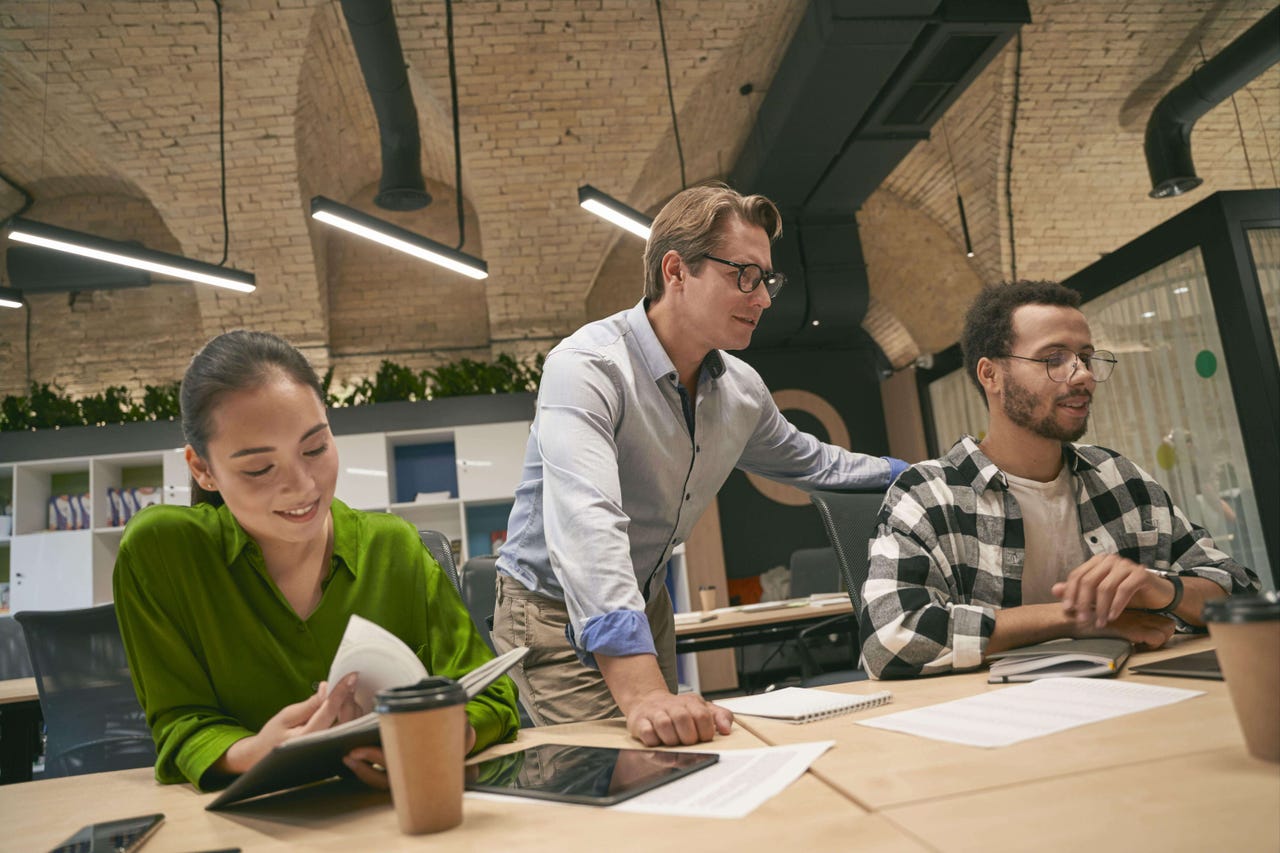 Man standing next to two seated coworkers who are working on a project together. They all look happy.